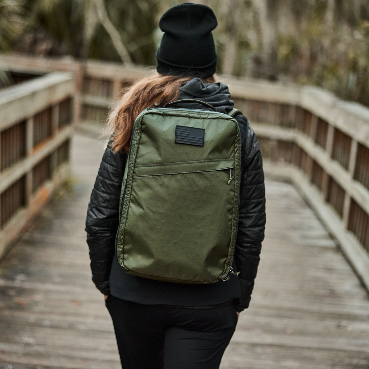 Dressed in a black outfit and beanie, a person strolls across a wooden bridge with the GR1 USA - X-PAC, featuring AquaGuard zippers for added protection against the elements.