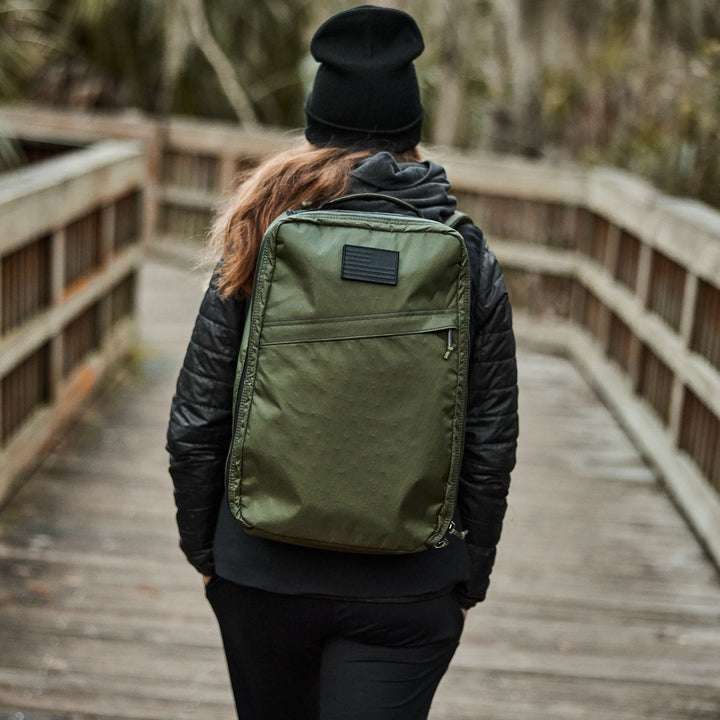 Dressed in a black outfit and beanie, a person strolls across a wooden bridge with the GR1 USA - X-PAC, featuring AquaGuard zippers for added protection against the elements.