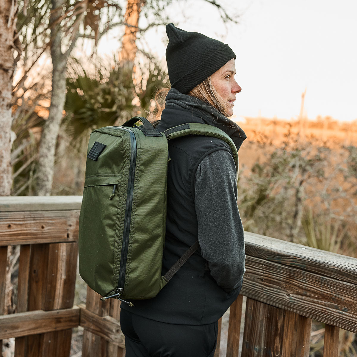 A person with a green GR1 USA - X-PAC backpack and black beanie looks over a wooden railing in nature, their gear set for anything with AquaGuard zippers.