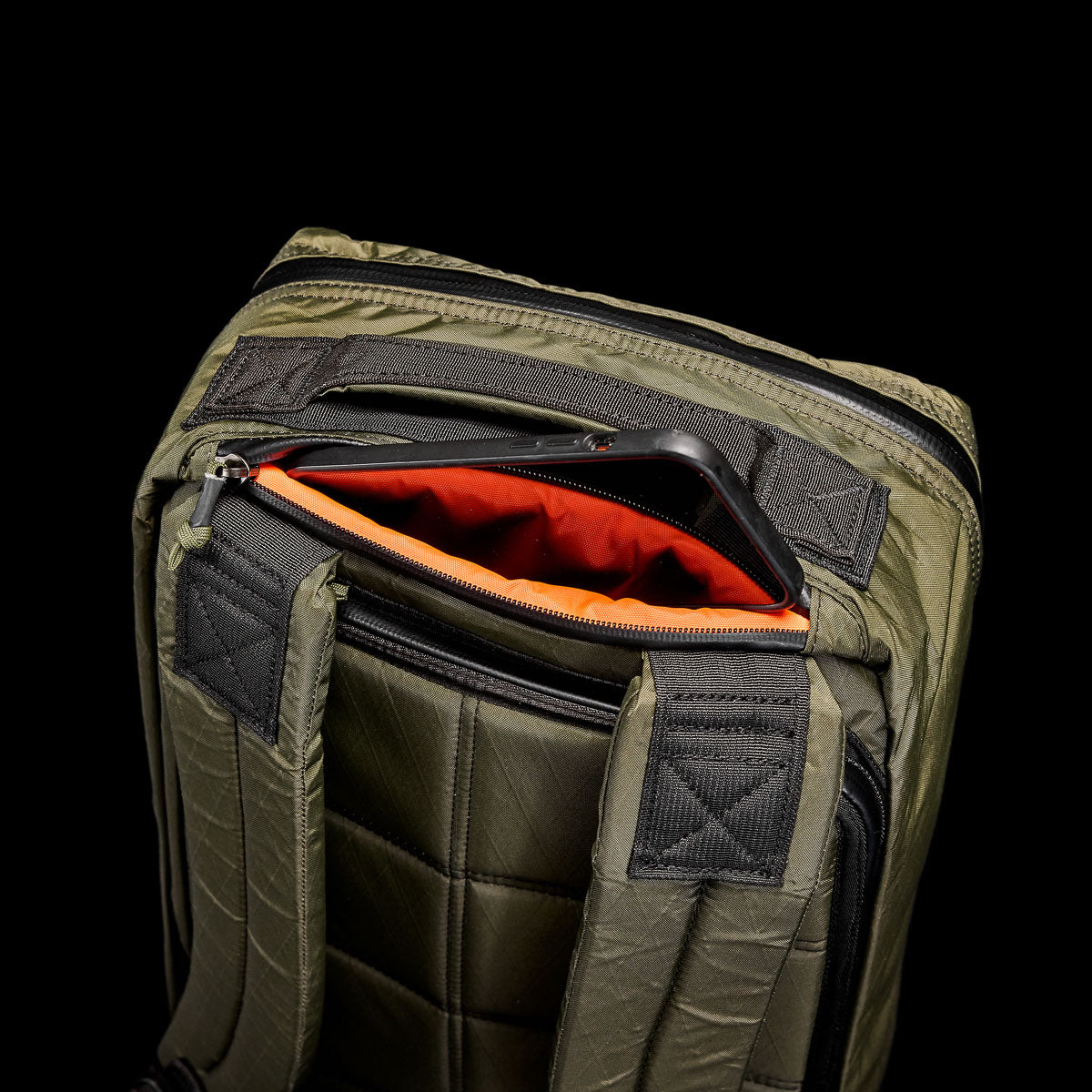 The Olive GR1 USA - X-PAC backpack with AquaGuard zippers displays a partially opened red-lined pocket, revealing a black smartphone inside.