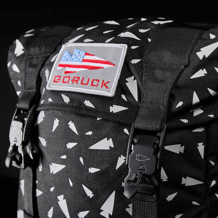 Close-up of the GORUCK M23 Spearhead Shrapnel rucksack featuring a black design with white geometric patterns. The front flap displays a bold patch of the American flag along with the "GORUCK" text. This bag is crafted from durable Cordura material and includes black buckles to secure the top.