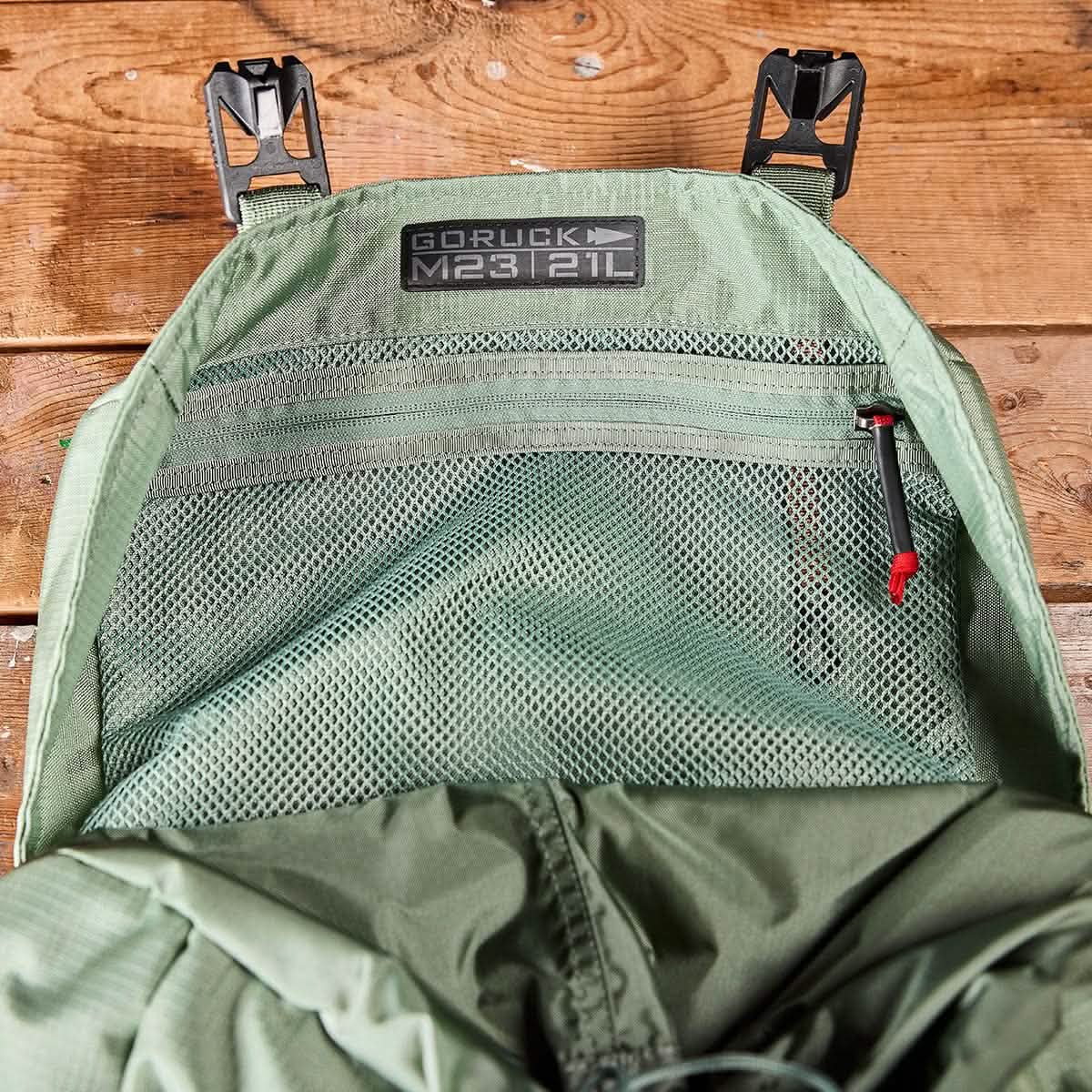 Close-up of an open Seaspray + Speed Tan pack, showcasing the inner mesh pocket with a zipper. The label on the interior reads GORUCK M23 - Ripstop ROBIC® 21L, highlighting its use of durable materials. The background is a wooden surface.