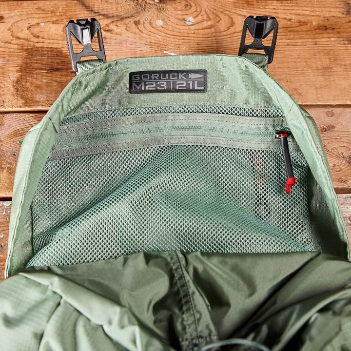 Close-up of an open Seaspray + Speed Tan pack, showcasing the inner mesh pocket with a zipper. The label on the interior reads GORUCK M23 - Ripstop ROBIC® 21L, highlighting its use of durable materials. The background is a wooden surface.