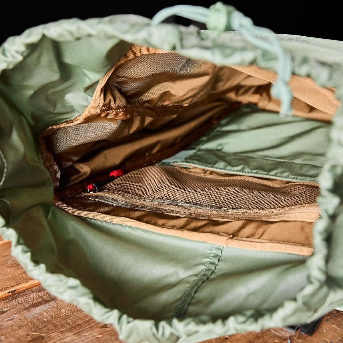 A GORUCK M23 rucksack in Seaspray with a Speed Tan interior lining rests on a wooden surface. Made from durable Ripstop ROBIC® materials, it features a mesh pocket with a red zipper that offers a glimpse of its compartmentalized storage design.