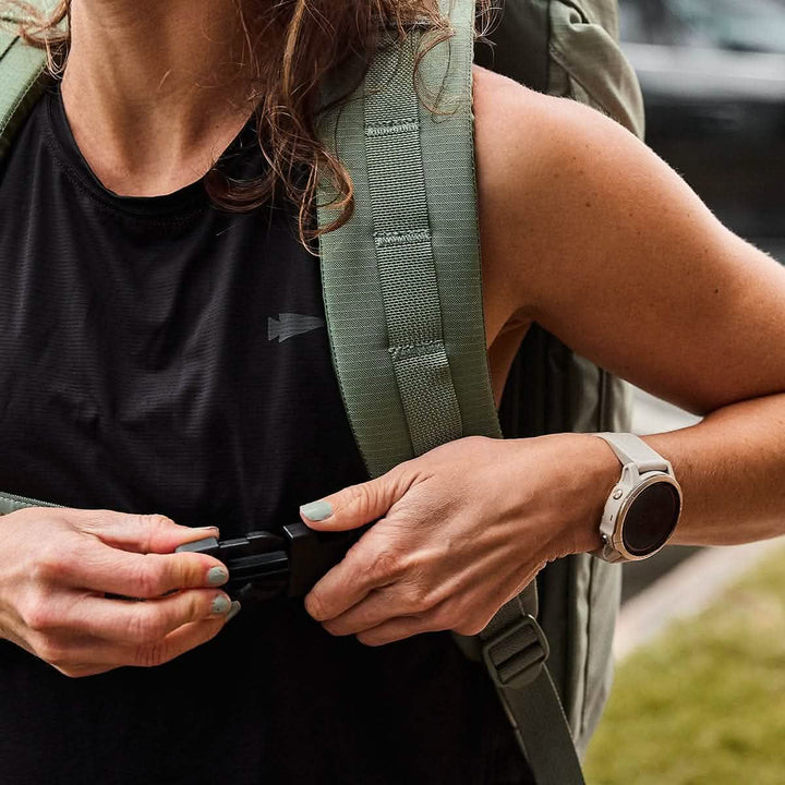 A person in a black sleeveless top adjusts the strap of their GORUCK M23 - Ripstop ROBIC® rucksack in Seaspray + Speed Tan. They wear a smartwatch on their wrist and are outdoors, with a blurred background featuring a vehicle. The rucksack appears to be constructed from durable materials, ideal for the journey ahead.
