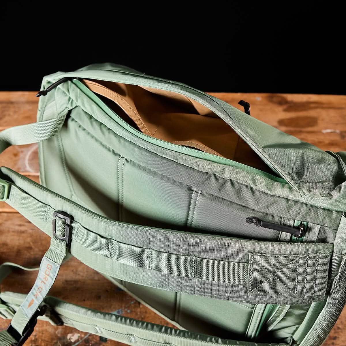 The M23 - Ripstop ROBIC® - Seaspray + Speed Tan messenger bag by GORUCK, made from robust materials, sits on a wooden surface, revealing its speed tan interior. It is equipped with a padded shoulder strap and includes multiple compartments along with a back pocket secured by a zipper.