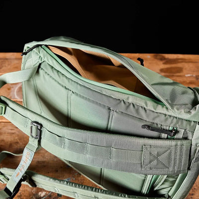 The M23 - Ripstop ROBIC® - Seaspray + Speed Tan messenger bag by GORUCK, made from robust materials, sits on a wooden surface, revealing its speed tan interior. It is equipped with a padded shoulder strap and includes multiple compartments along with a back pocket secured by a zipper.