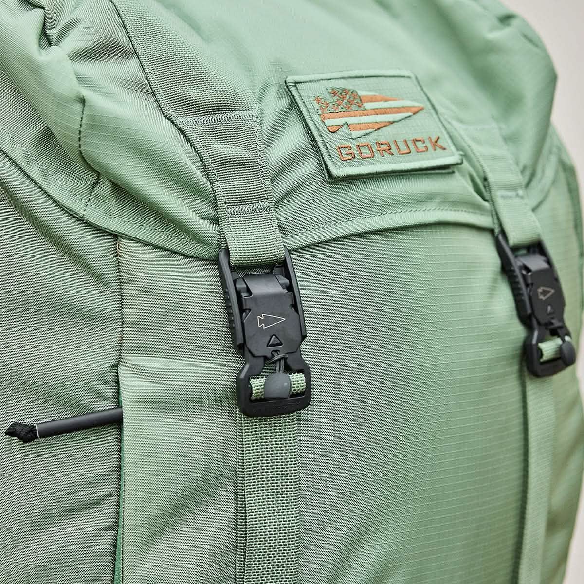 Close-up of the M23 - Ripstop ROBIC® pack in Seaspray and Speed Tan by GORUCK, showcasing its durable materials and textured fabric. The rucksack features black buckles and straps, with a patch displaying the GORUCK logo—an arrow and stripes design—sewn on the top flap.