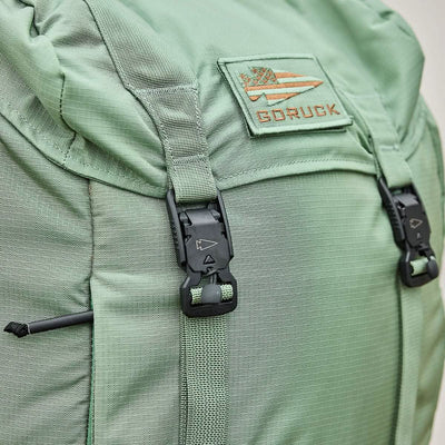 Close-up of the M23 - Ripstop ROBIC® pack in Seaspray and Speed Tan by GORUCK, showcasing its durable materials and textured fabric. The rucksack features black buckles and straps, with a patch displaying the GORUCK logo—an arrow and stripes design—sewn on the top flap.