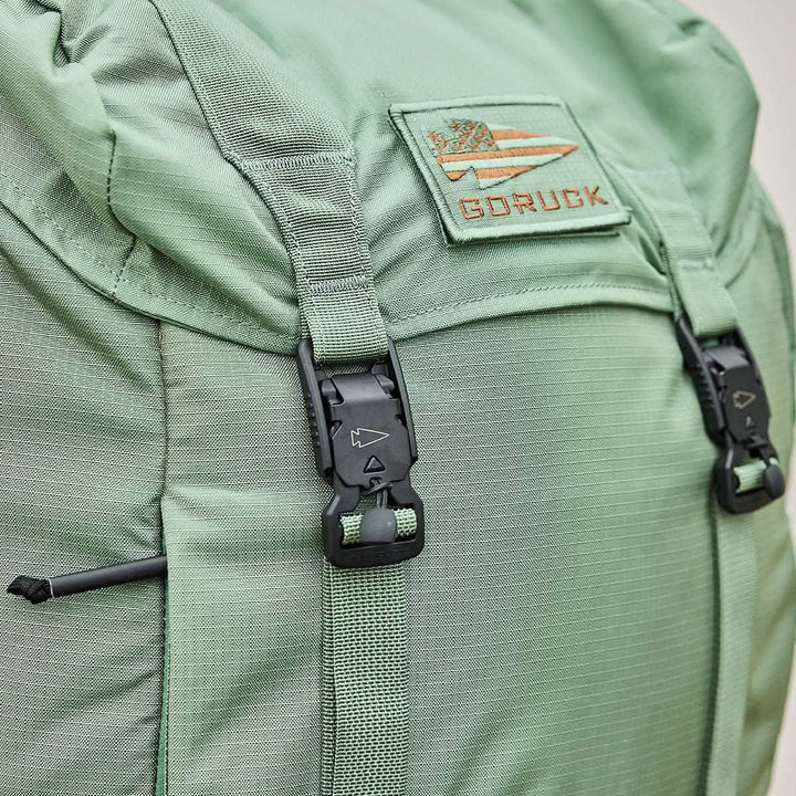 Close-up of the M23 - Ripstop ROBIC® pack in Seaspray and Speed Tan by GORUCK, showcasing its durable materials and textured fabric. The rucksack features black buckles and straps, with a patch displaying the GORUCK logo—an arrow and stripes design—sewn on the top flap.