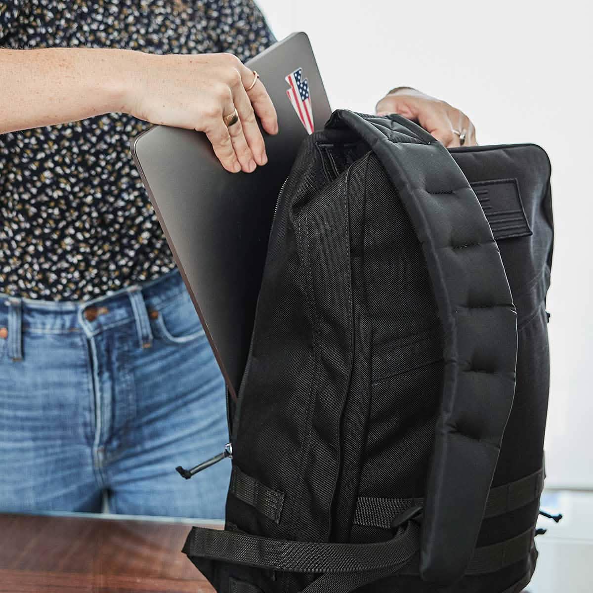 A person carefully places a laptop adorned with an American Manufacturing flag sticker into the sleek GR1 USA - Cordura backpack by GORUCK, ready for a mission that seems almost Special Forces worthy.