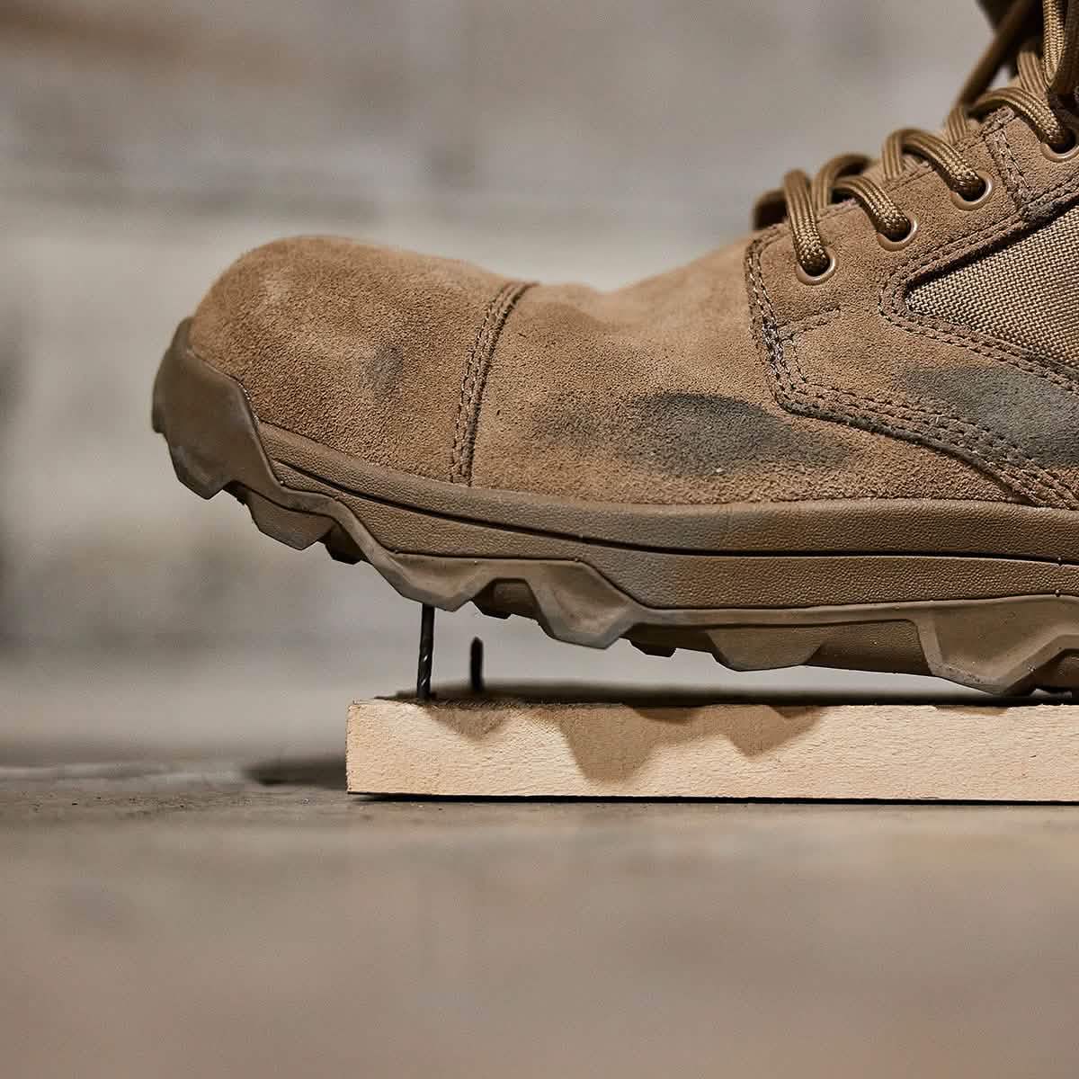 Zoomed-in perspective of a tan suede MACV-2 Safety Boot - Mid Top by GORUCK, featuring rugged tread on a wooden surface. The untied laces and blurred background accentuate the texture and intricate details of this versatile safety boot.