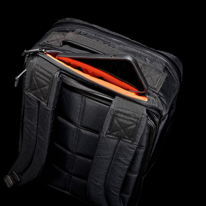 The GR1 USA - X-PAC backpack boasts an open top compartment unveiling an orange interior with a partially visible tablet. Made from waterproof materials and featuring AquaGuard zippers, it provides stylish protection for your essentials.