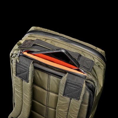 An olive green GR1 USA - X-PAC rucksack, showcasing a tablet in the top pocket, designed from durable X-PAC material against a black backdrop.