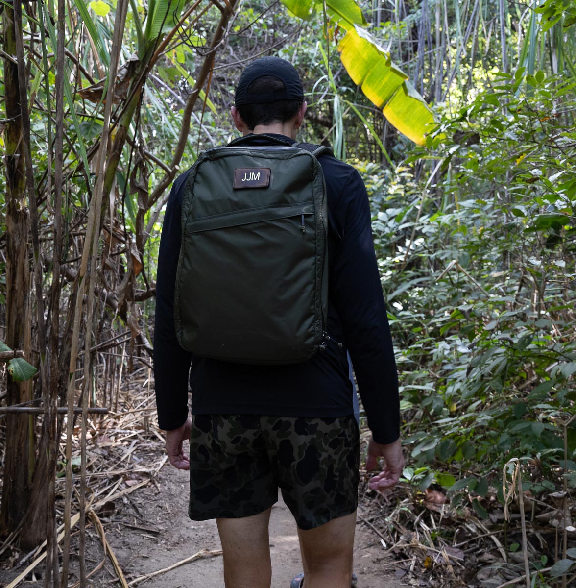 Wearing a GR1 USA - X-PAC waterproof rucksack, an individual strides along a forest trail, surrounded by dense green foliage.
