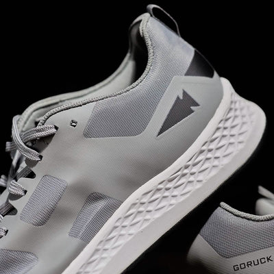 A close-up of the GORUCK Women's Rough Runner - Speed Grey + White athletic shoes, showcasing their EVA midsole with a textured white sole and geometric side design. The shoes are set against a black background, accentuating their modern and sleek appearance.