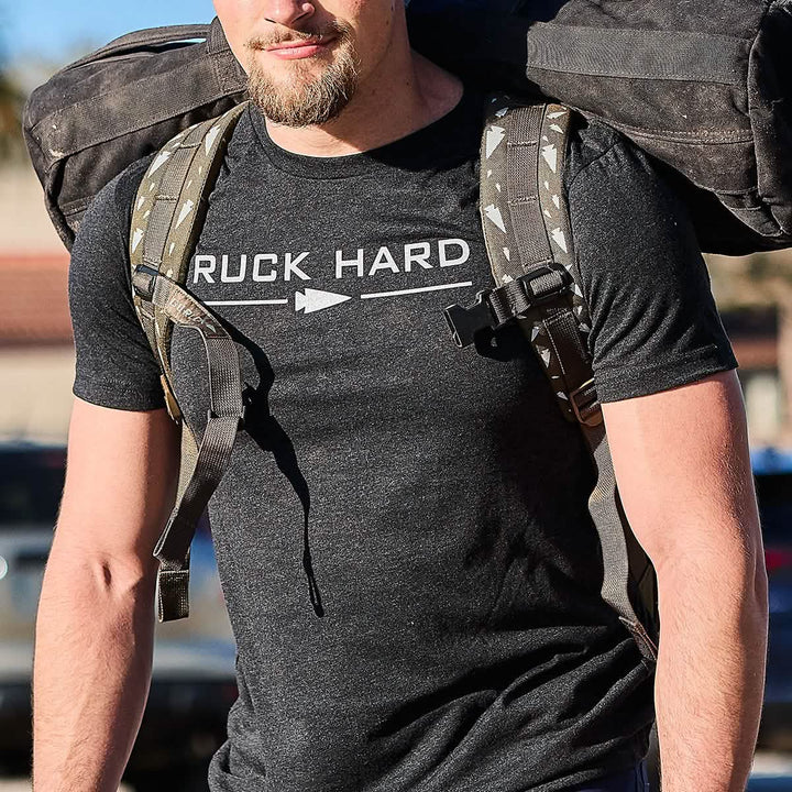 A fitness enthusiast walks through the sunny outdoors with a large backpack, wearing his "Ruck Hard Tee - Tri-Blend," symbolizing his dedication to training and passion for the ruck lifestyle.