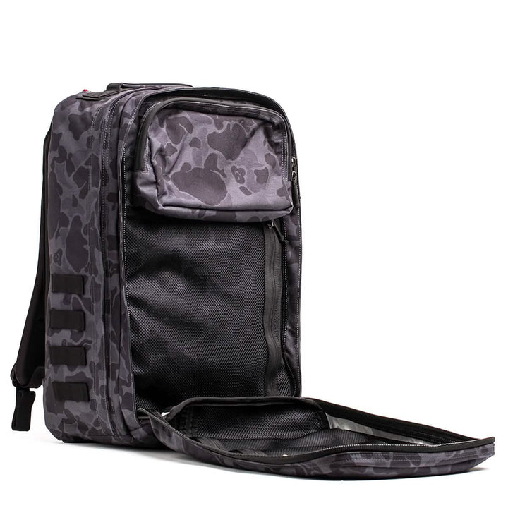 The GORUCK GR2 backpack, in a black and gray camo pattern reminiscent of Special Forces gear, stands upright with zippers partially open. It reveals a spacious inner compartment with a mesh pocket and features padded shoulder straps along with a sturdy handle on top.
