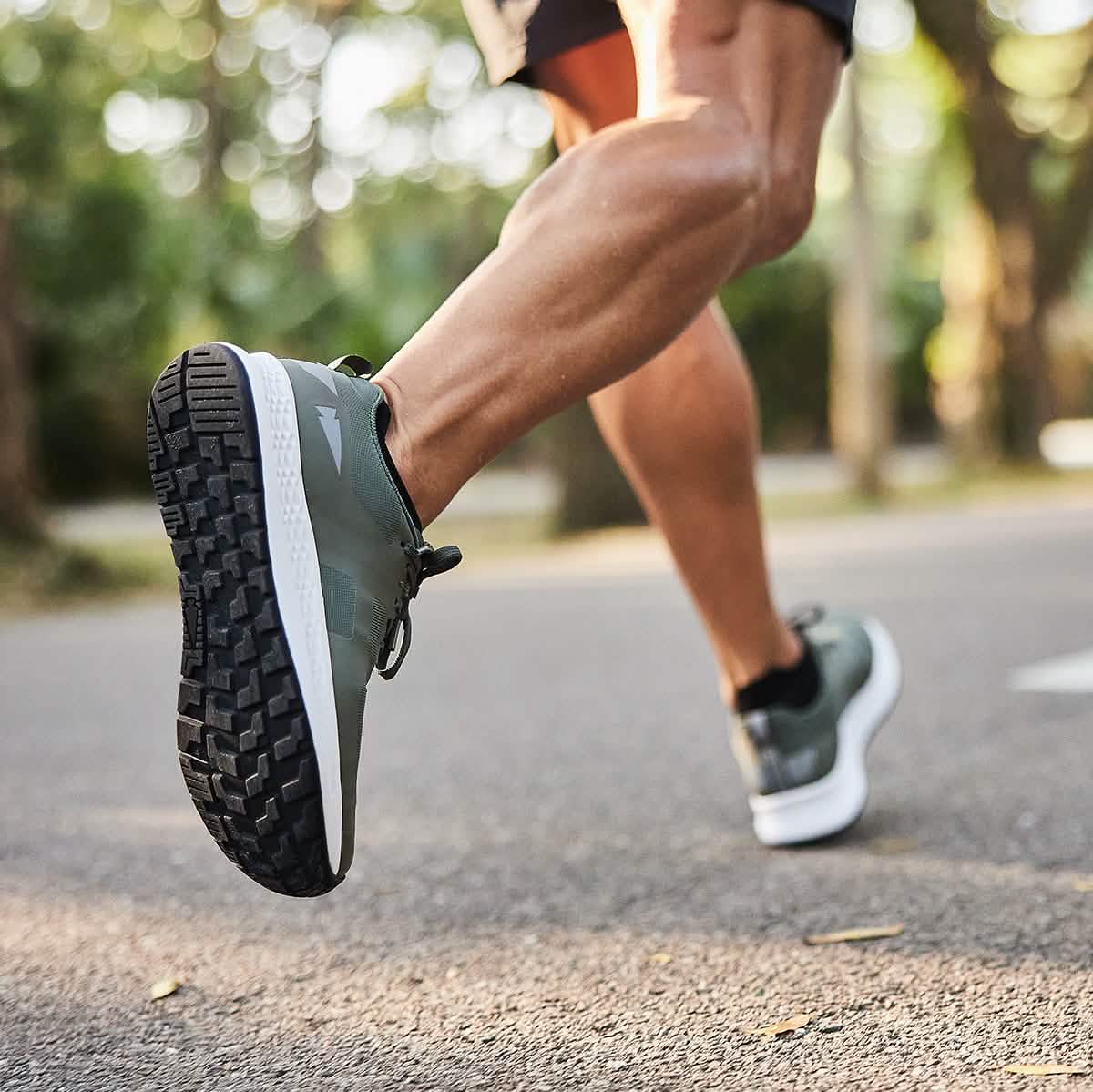 A person running on a paved path in a park showcases their Men's Rough Runner - Earth shoes by GORUCK, accentuating their muscular legs. The scene features trees and a sunny atmosphere, emphasizing the gradient density midsole of the shoes that makes them ideal for varied terrains.