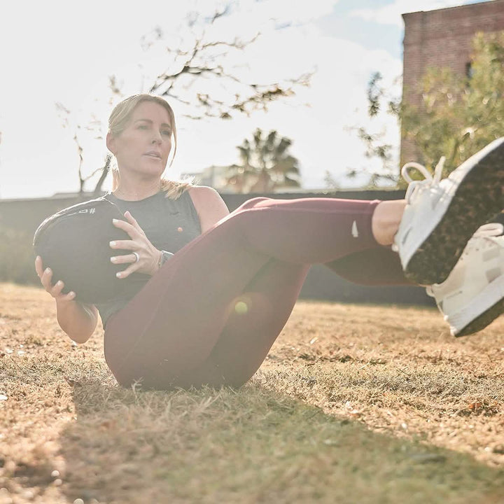 Exercising outdoors on the grass, a person holds a Sand Medicine Ball by GORUCK, twisting to enhance core stability. Under the bright sunlight casting a warm glow, they sport a sleeveless top, maroon leggings, and white sneakers.