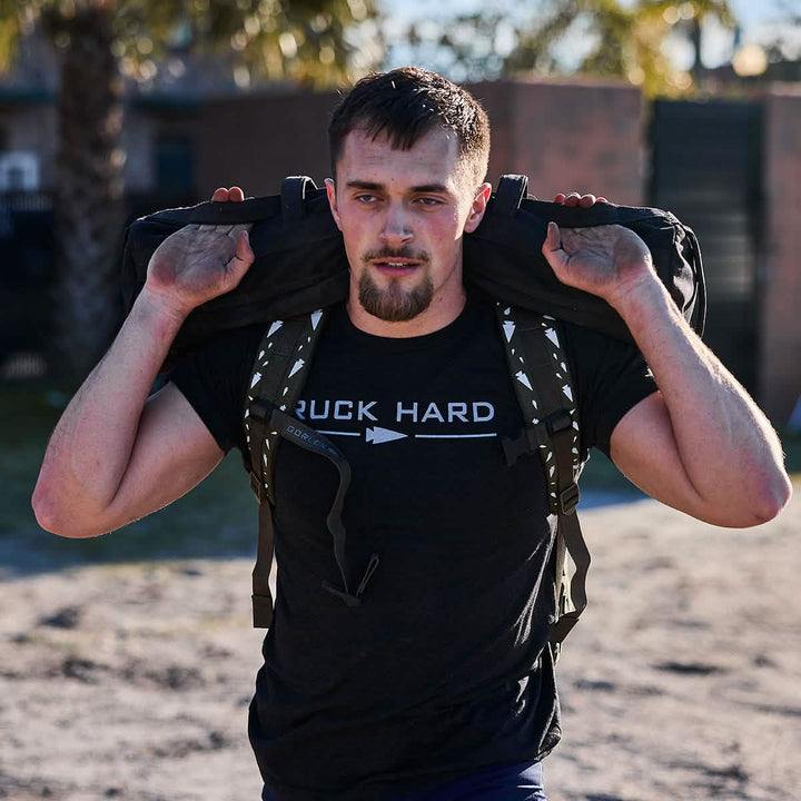A person embracing their fitness journey strides outdoors with a heavy backpack and proudly wears a "Ruck Hard Tee - Tri-Blend," embodying the spirit of intense training.