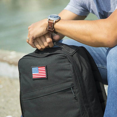 A person wearing a watch and jeans sits outdoors, resting their arm on a black backpack that features an embroidered Patch - American Flag by vendor-unknown. The background is blurred, indicating a natural setting.