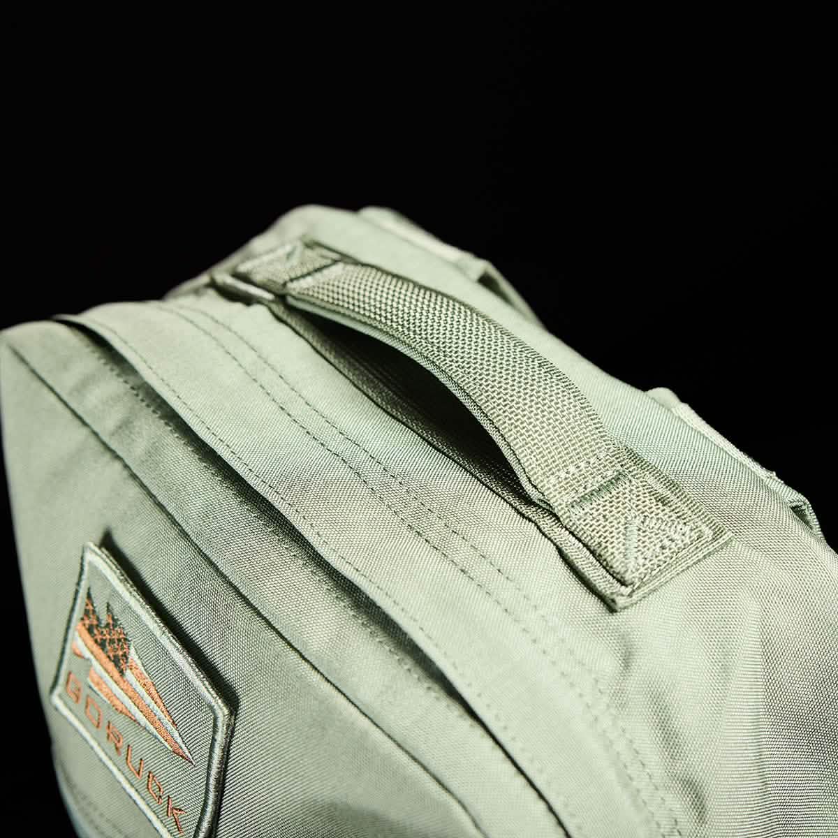 Close-up of the KR1 kids rucksack by GORUCK in green fabric, complete with a woven handle and padded straps. It includes a patch featuring triangular designs and the text "GORUCK" in orange. The black background highlights the bag's details, making it perfect for carrying school supplies.