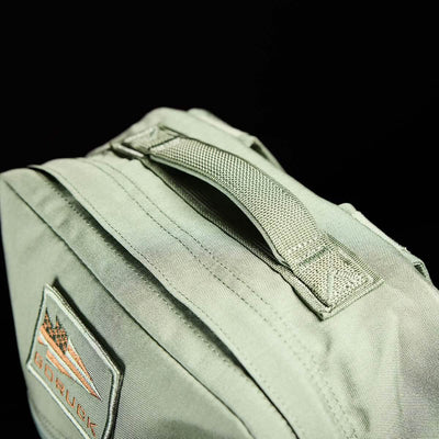 Close-up of the KR1 kids rucksack by GORUCK in green fabric, complete with a woven handle and padded straps. It includes a patch featuring triangular designs and the text "GORUCK" in orange. The black background highlights the bag's details, making it perfect for carrying school supplies.