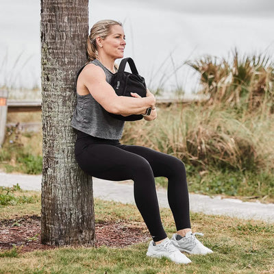 A woman engages in the GORUCK Kettle Bell Curve Training Program with Nichele, performing a seated wall squat against a palm tree while holding a weight bag. She is dressed in a gray sleeveless top, black leggings, and white sneakers as she completes her full-body workout outdoors on a cloudy day amidst grassy surroundings.