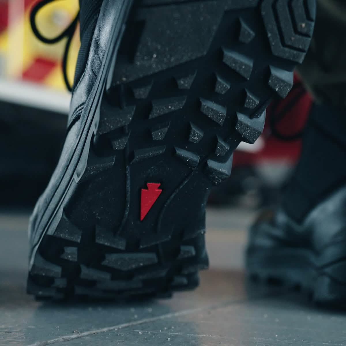 A close-up shot of the MACV-2 Safety Boot - High Top by GORUCK is depicted, showcasing a black safety boot with rugged tread. It meets Special Forces standards and features a distinctive red symbol in the center. The sole is engineered for traction, as demonstrated by its thick, angular patterns. The background is slightly out of focus, adding a hint of color to the scene.