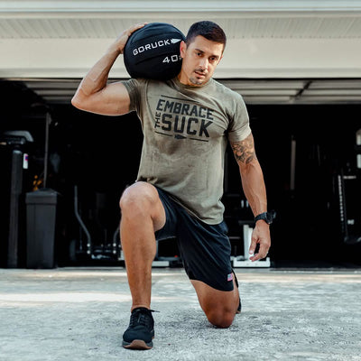 A man kneels on one leg in the driveway, with a GORUCK Sand Medicine Ball resting on his shoulder. He's dressed in a tan "Embrace the Suck" t-shirt and dark shorts, with a garage behind him filled with core stability equipment, highlighting his well-equipped home gym.