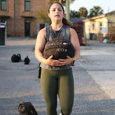 A person in a gray tank top and green leggings demonstrates core stability outdoors by carrying a GORUCK Sand Medicine Ball weighing 40 pounds, with buildings and kettlebells in the background that resemble an open-air extension of a home gym.