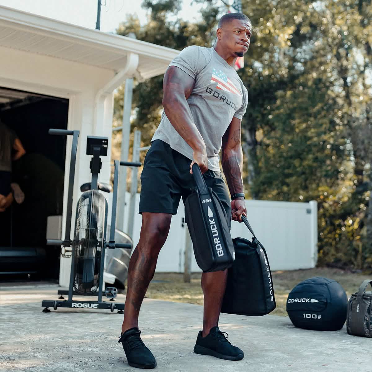 A person is exercising outdoors with fitness equipment, lifting a Sand Jerry Can in one hand to build core strength. They are wearing a gray GORUCK shirt and black shorts. Various weights and a fitness bike are in the background, resembling gear made from Special Forces Grade materials. Trees and a white fence are visible.
