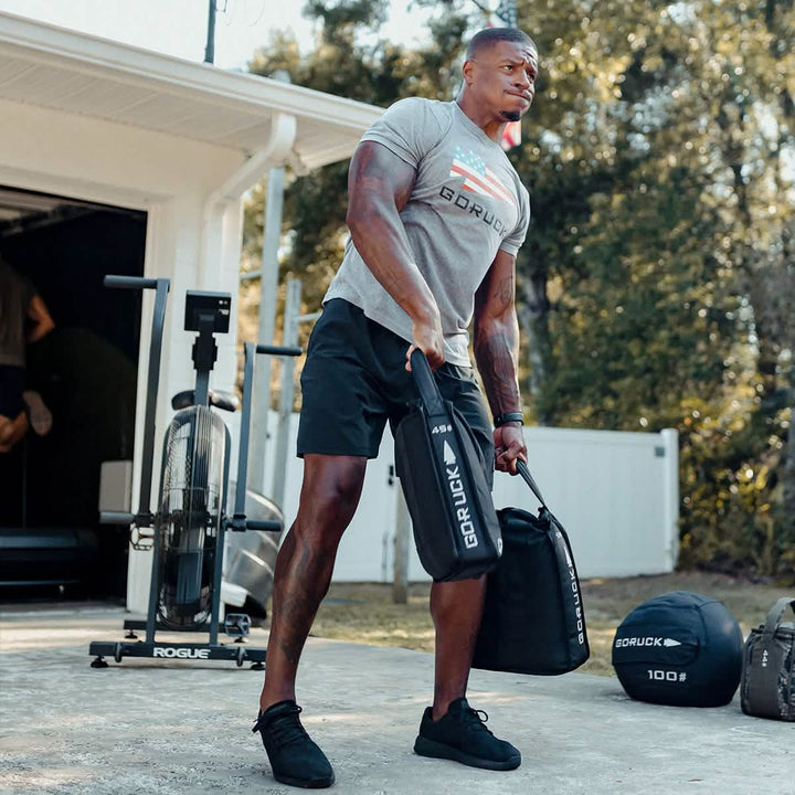 A person is exercising outdoors with fitness equipment, lifting a Sand Jerry Can in one hand to build core strength. They are wearing a gray GORUCK shirt and black shorts. Various weights and a fitness bike are in the background, resembling gear made from Special Forces Grade materials. Trees and a white fence are visible.