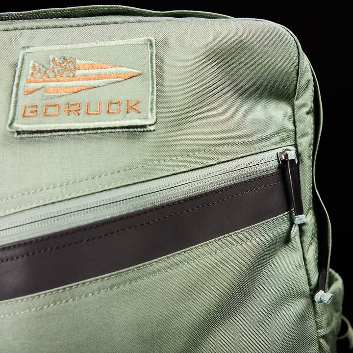 A detailed view of the green KR1 kids rucksack from GORUCK showcases a branded patch near the top. This backpack includes padded straps for comfort and features a front horizontal zipper accentuated by a dark brown strip, all against a black backdrop. It's ideal for stylishly carrying school supplies.