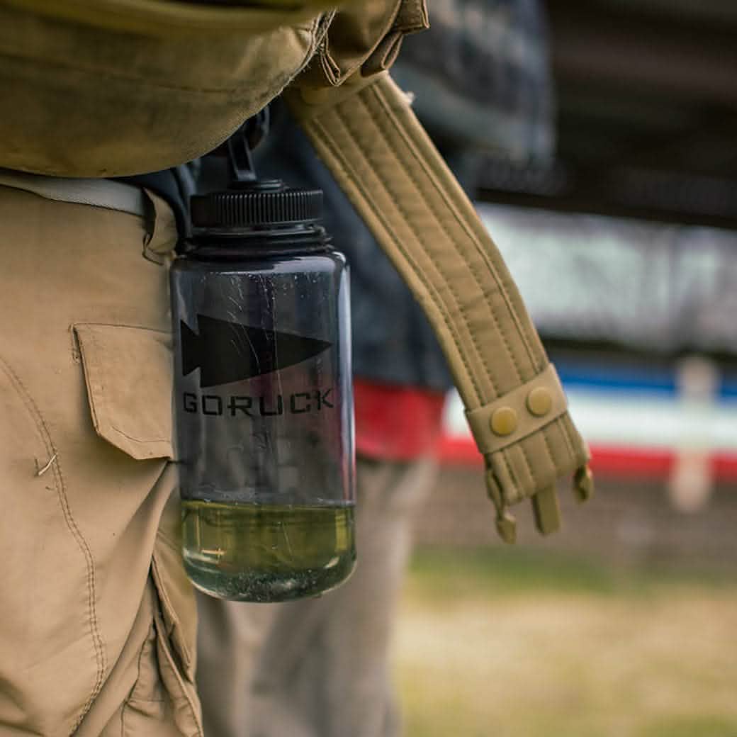 Close-up of a person wearing tan pants with a large, leak-proof GORUCK Spearhead Nalgene Water Bottle (32oz) secured to a strap. The bottle is partially filled with liquid. The blurred background suggests another person and an outdoor setting, hinting at an adventure on the horizon.