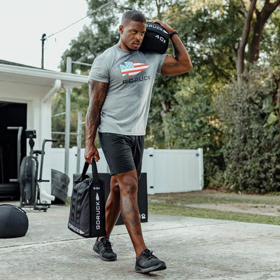 A man in athletic clothing carries a GORUCK Sand Jerry Can over his shoulder while holding a black fitness bag crafted with Special Forces Grade materials in the other hand. He strides along a concrete path near a garage, surrounded by trees and gym equipment, enhancing his core strength with each determined step.
