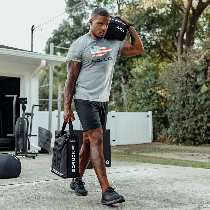 A man in athletic clothing carries a GORUCK Sand Jerry Can over his shoulder while holding a black fitness bag crafted with Special Forces Grade materials in the other hand. He strides along a concrete path near a garage, surrounded by trees and gym equipment, enhancing his core strength with each determined step.