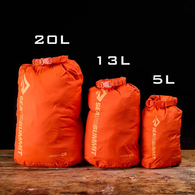 Three orange Sea to Summit Dry Sacks in sizes 20L, 13L, and 5L are positioned on a wooden surface against a black background. Each sack is made from lightweight nylon and stands upright, showcasing its roll-top waterproof closure and size labels.