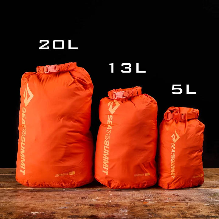 Three orange Sea to Summit Dry Sacks in sizes 20L, 13L, and 5L are positioned on a wooden surface against a black background. Each sack is made from lightweight nylon and stands upright, showcasing its roll-top waterproof closure and size labels.