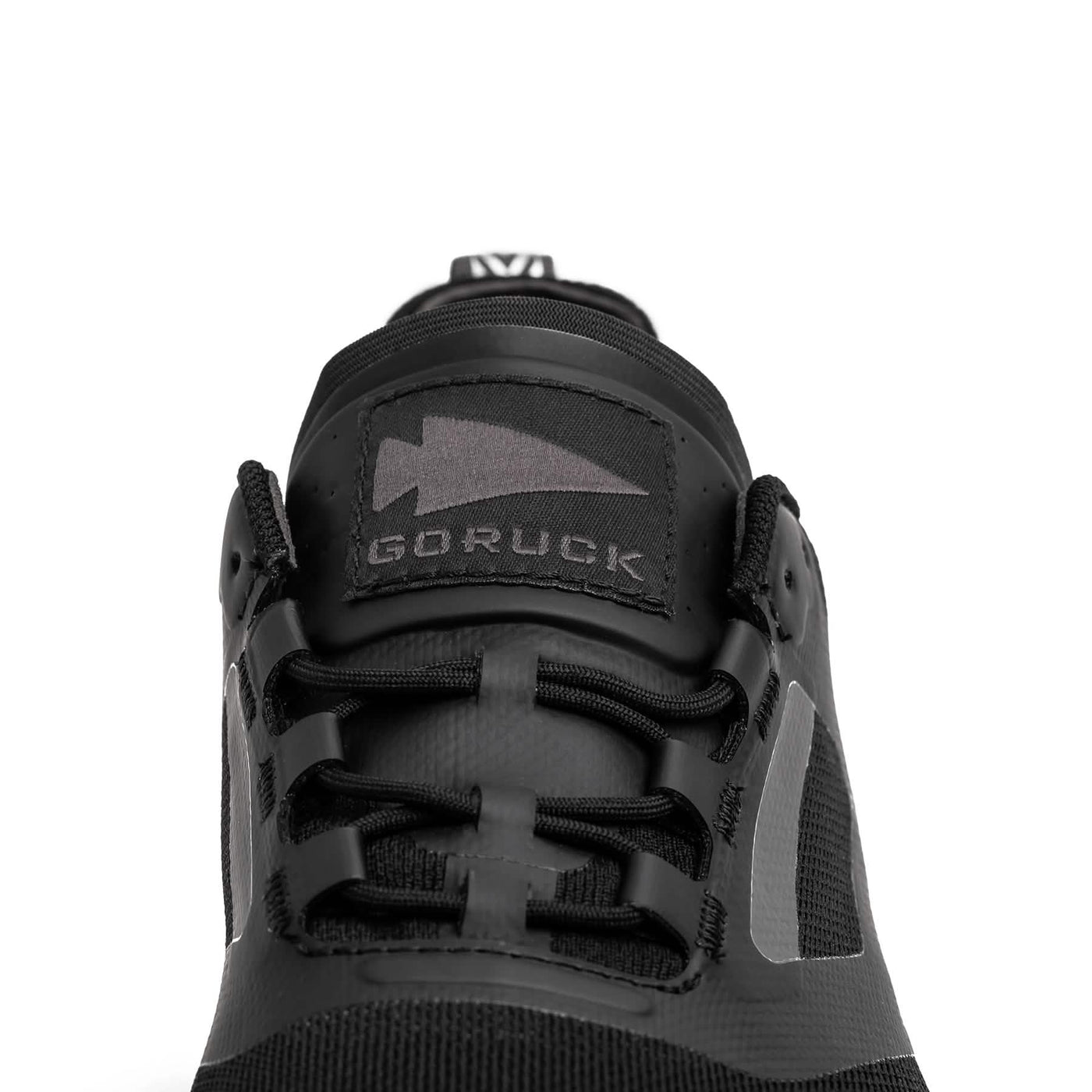 Close-up of a black athletic shoe from GORUCK, featuring the Mackall - Black + Forged Iron design. This sleek trail shoe has black laces and a prominent arrow logo, embodying the durability required for Special Forces training.