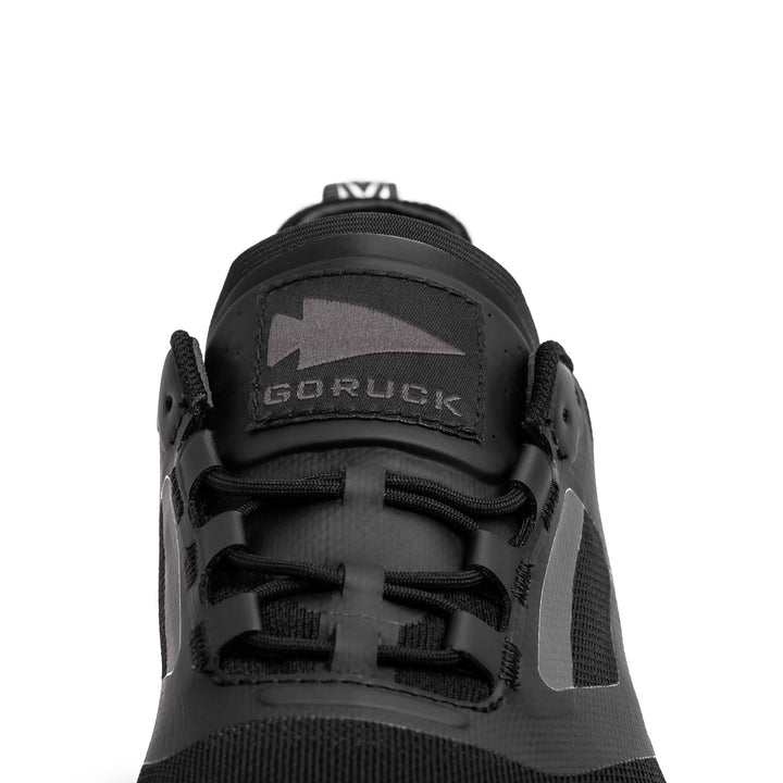 Close-up of a black athletic shoe from GORUCK, featuring the Mackall - Black + Forged Iron design. This sleek trail shoe has black laces and a prominent arrow logo, embodying the durability required for Special Forces training.