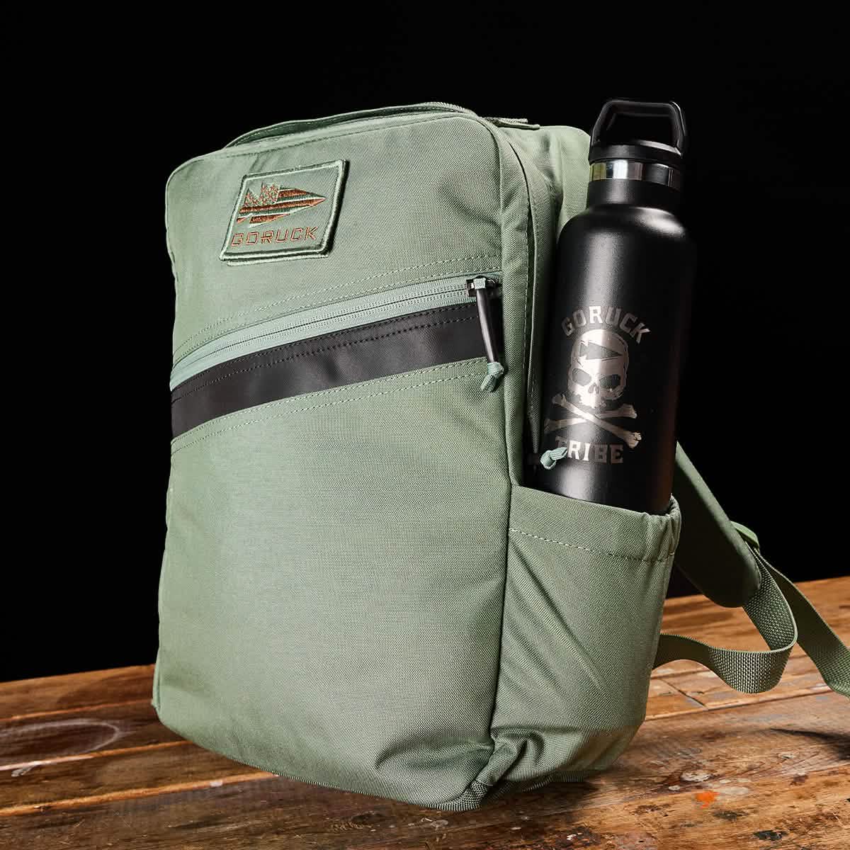 The GORUCK KR1 green kids' rucksack, featuring padded straps and a front zipper pocket with a top patch, holds a black water bottle adorned with a skull and text in its side pocket. This backpack, ideal for school supplies, is placed on a wooden surface against a black backdrop.