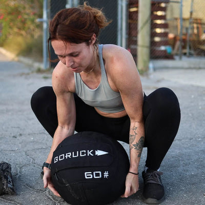 A person crouches on a concrete path, grasping a GORUCK Sand Medicine Ball weighing 60 pounds, ideal for enhancing core stability. They are dressed in a gray sports bra and black leggings. Behind them, a chain-link fence and assorted materials suggest an environment suitable for a home gym.