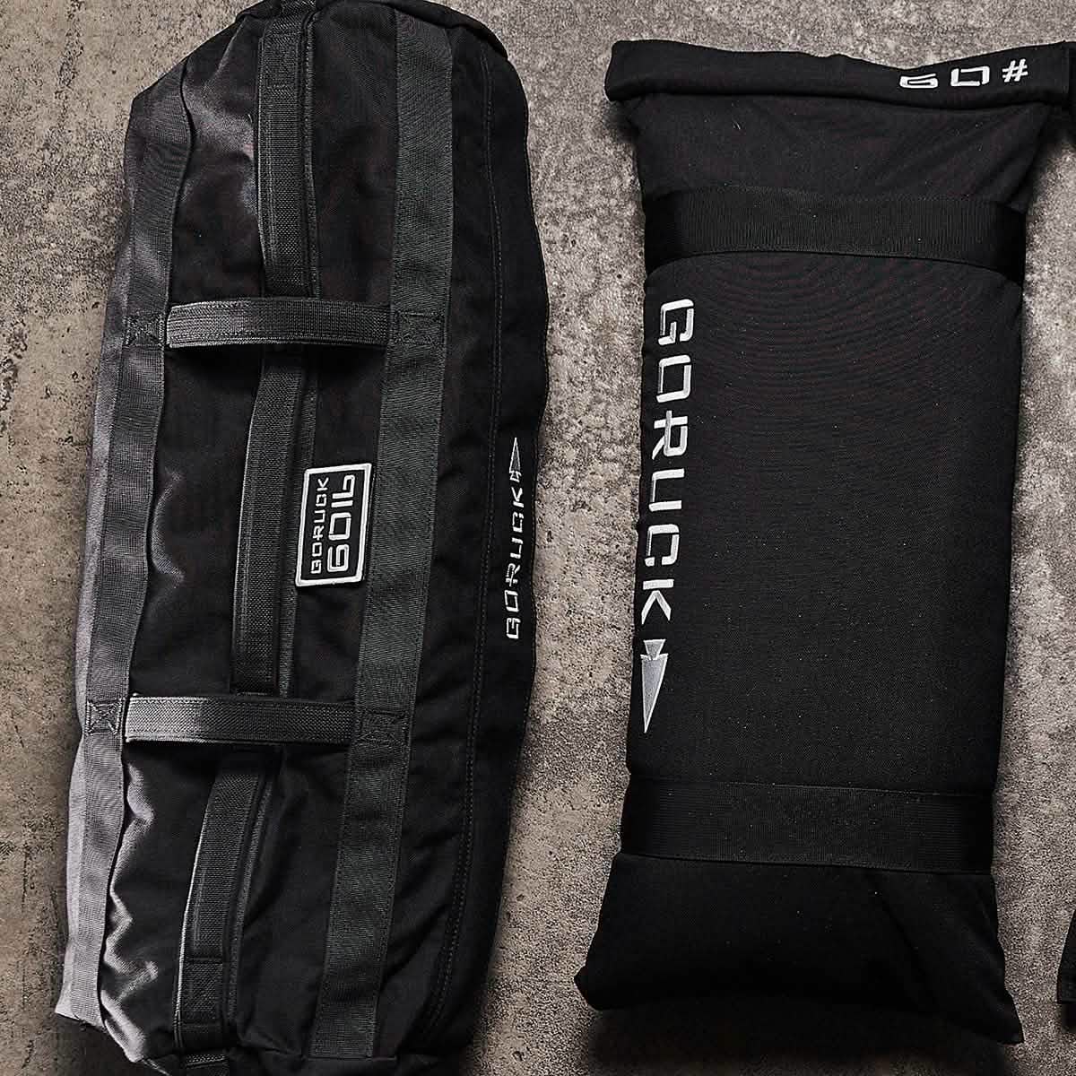 Two Sandbags 2.0 by GORUCK, crafted from durable black 1000D CORDURA and featuring white logos, are placed on a textured surface. The larger training sandbag with multiple handles is positioned on the left, while the smaller one lies flat on the right. Both are backed by the trusted Scars Lifetime Guarantee.