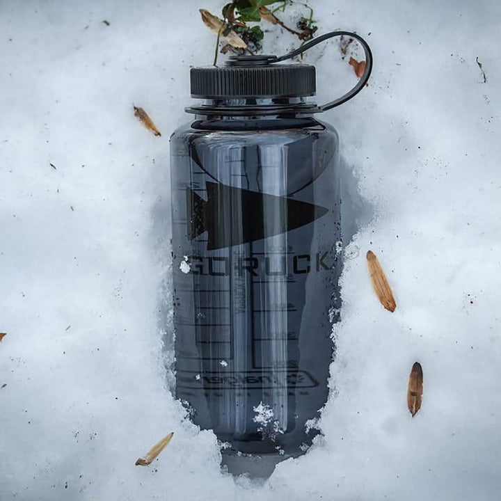 A durable Nalgene Water Bottle (32oz) - GORUCK Spearhead is half-buried in the snow, with twigs and small plants scattered around it. The bottle features a black screw-on lid and a handle loop, ready for your next adventure.