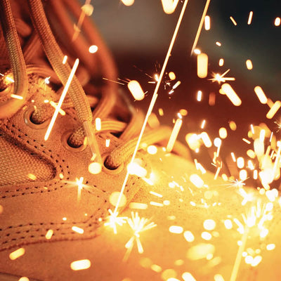 Close-up of tan GORUCK MACV-2 Safety Boot - Mid Top with large sparks flying around them, suggesting metal welding or grinding. The bright orange and yellow sparks contrast with the textured surface of the boots, creating a dynamic and energetic scene.