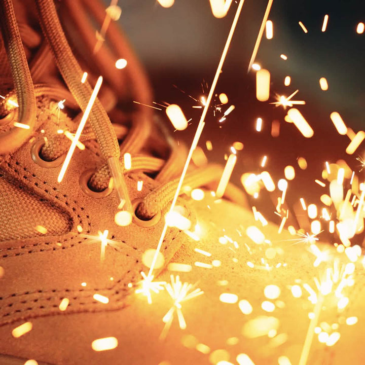 Close-up of tan GORUCK MACV-2 Safety Boot - Mid Top with large sparks flying around them, suggesting metal welding or grinding. The bright orange and yellow sparks contrast with the textured surface of the boots, creating a dynamic and energetic scene.