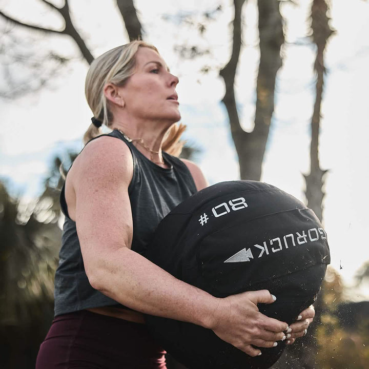 A woman wearing a sleeveless top exercises outdoors with the GORUCK Sand Medicine Ball, enhancing her core stability. She looks focused amid the trees, showcasing a workout that's perfect for a home gym.