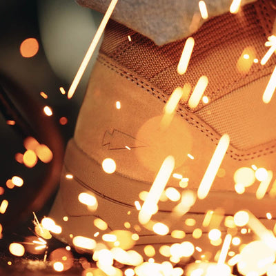 A close-up captures someone in the MACV-2 Safety Boot - Mid Top by GORUCK, made from suede and emitting orange sparks. The heel showcases a subtle embossed lightning bolt design, adhering to Special Forces Standards, hinting at a busy workshop or construction environment with active metal grinding.